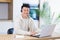 Young asian man with headset looks at camera and smiles sitting at home in the kitchen and working on laptop