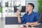 Young Asian man holding a small plastic fan blowing to his face because of hot weather while sitting in a chair in the office.