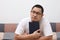 Young Asian man looking up and thinking while holding book and sitting on cozy sofa in apartment