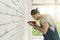 Young Asian man using electric drill on white brick wall in room