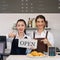 Young asian shopkeeper and  caucasian barista with a smile holds an OPEN sign in front of a coffee shop counter. Morning