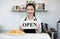 Young asian shopkeeper with a smile holds an OPEN sign in front of a coffee shop counter. Morning atmosphere in a coffee shop