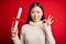 Young asian student woman holding graduate diploma over red isolated background doing ok sign with fingers, excellent symbol