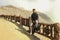 A young Asian tourist sits on a fence at the edge of Kawah Ijen crater. Man on edge of crater with colorful sky at