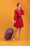 Young Asian woman bring suitcase and using phone isolated over orange background
