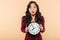 Young asian woman with curly long hair holding clock showing nearly 8 being late or missing something over peach background