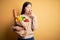 Young asian woman holding paper bag of fresh healthy groceries over yellow isolated background bored yawning tired covering mouth