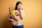 Young asian woman holding paper bag of fresh healthy groceries over yellow isolated background Yawning tired covering half face,