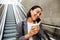 Young asian woman talking on cellphone and drinking coffee while standing on escalator