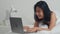 Young Asian woman using laptop checking social media feeling happy smiling while lying on bed after wake up at house in the