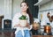 Young asian woman in white shirt hold basket with green house plant in wicker pot in hands on kitchen