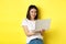 Young asian woman working freelance, using laptop and smiling, standing over yellow background