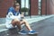 Young athlete in blue windrunner sitting on the street adjusting running program for his morning workout using smart watches on