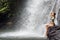 Young athletic woman with long hair sitting on stone on background of waterfall with many drops and tropical forest