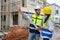 Young attractive construction man and woman in vests with helmets working on the under-construction building site. Home building