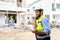 Young attractive construction smiling bearded using smartphone calling to client, worker in vest with white helmet standing on