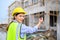 Young attractive construction woman in safety vest with yellow helmet working with radio, standing on building construction site.