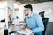 Young attractive dark-haired man in glassess is working with a computer at his workplace in office. He wears blue shirt