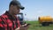 Young attractive farmer with phone standing in field, tractor working in green field in background. Smart farming using