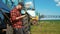 Young attractive farmer with phone standing in field, tractor working in green field in background. Smart farming using