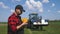 Young attractive farmer with phone standing in field, tractor working in green field in background. Smart farming using