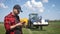 Young attractive farmer with phone standing in field, tractor working in green field in background. Smart farming using