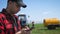 Young attractive farmer with phone standing in field, tractor working in green field in background. Smart farming using