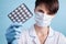 A young attractive female scientist in a mask holds a package of pills with gloved fingers on blue background