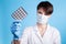 A young attractive female scientist in a mask holds a package of pills with gloved fingers on blue background