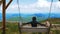 A young attractive female traveler rides a large swing with her arms outstretched to the sides at the top of the cliff