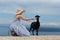 Young attractive female traveler wearing striped summer dress and straw hat squatting, feeding and petting black sheep