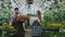 Young attractive florists couple in apron working in greenhouse. Cheerful man with box of flowers talking woman loosen