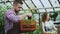 Young attractive florists couple in apron working in greenhouse. Cheerful man with box of flowers talking woman loosen