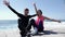 Young attractive happy couple raising the hands up in the air smiling and cheering sitting on the beach