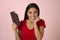 Young attractive and happy hispanic woman in red top smiling excited eating chocolate bar on pink background