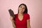 Young attractive and happy hispanic woman in red top smiling excited eating chocolate bar on pink background