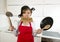 Young attractive home cook woman in red apron at kitchen holding pan and household with pot on her head in stress