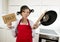 Young attractive home cook woman in red apron at kitchen holding pan and household with pot on her head in stress