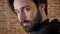 Young attractive man with beard turns head, watching at camera, smiling, flirt conception, brick background, side view