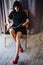 Young attractive woman with long legs in black elegant dress, sits in chair near window in interior of room