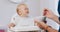 Young baby in white high baby chair and father's hands with a spoon and bowl, while he is feeding the baby, who