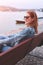 Young balanced redhead woman sitting on bench at riverbank side view in sunset