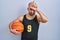 Young bald man with beard wearing basketball uniform holding ball making fun of people with fingers on forehead doing loser