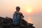 A young and bald man sits on top of a mountain in the background of the sea and dusk or sunset.