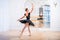 Young ballerina in a black tutu stands in graceful pose on pointe shoes in a large bright hall in front of a mirror
