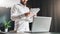 Young bearded atrractive businessman in white shirt is standing near desk in front of laptop, reading documents.