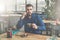 Young bearded attractive businessman in blue shirt is sitting at wooden table in restaurant, talking on phone