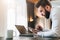 Young bearded businessman in shirt sitting at desk in front of laptop, making notes in digital tablet.