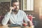 Young bearded businessman sits in cafe at table, drinking tea, talking on cellphone. On table notebook, pen.