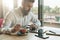 Young bearded businessman sits in office at table,uses tablet computer, drinks coffee.On desk is notebook,smartphone.